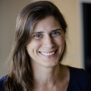 Person with long brown hair wearing a dark top, posed indoors against a softly lit background with neutral tones.