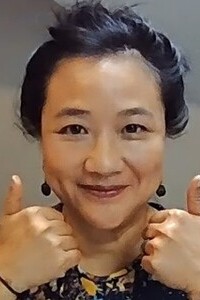 Two hands giving thumbs-up gesture, with dark hair tied back and wearing a patterned top.