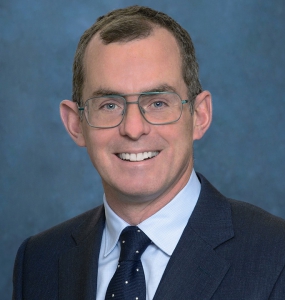 Person wearing a dark suit, white shirt, and navy polka-dot tie against a blue background.