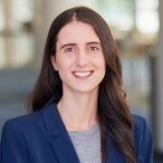 A person with long dark hair, wearing a blue blazer and gray top, smiles in an indoor setting.