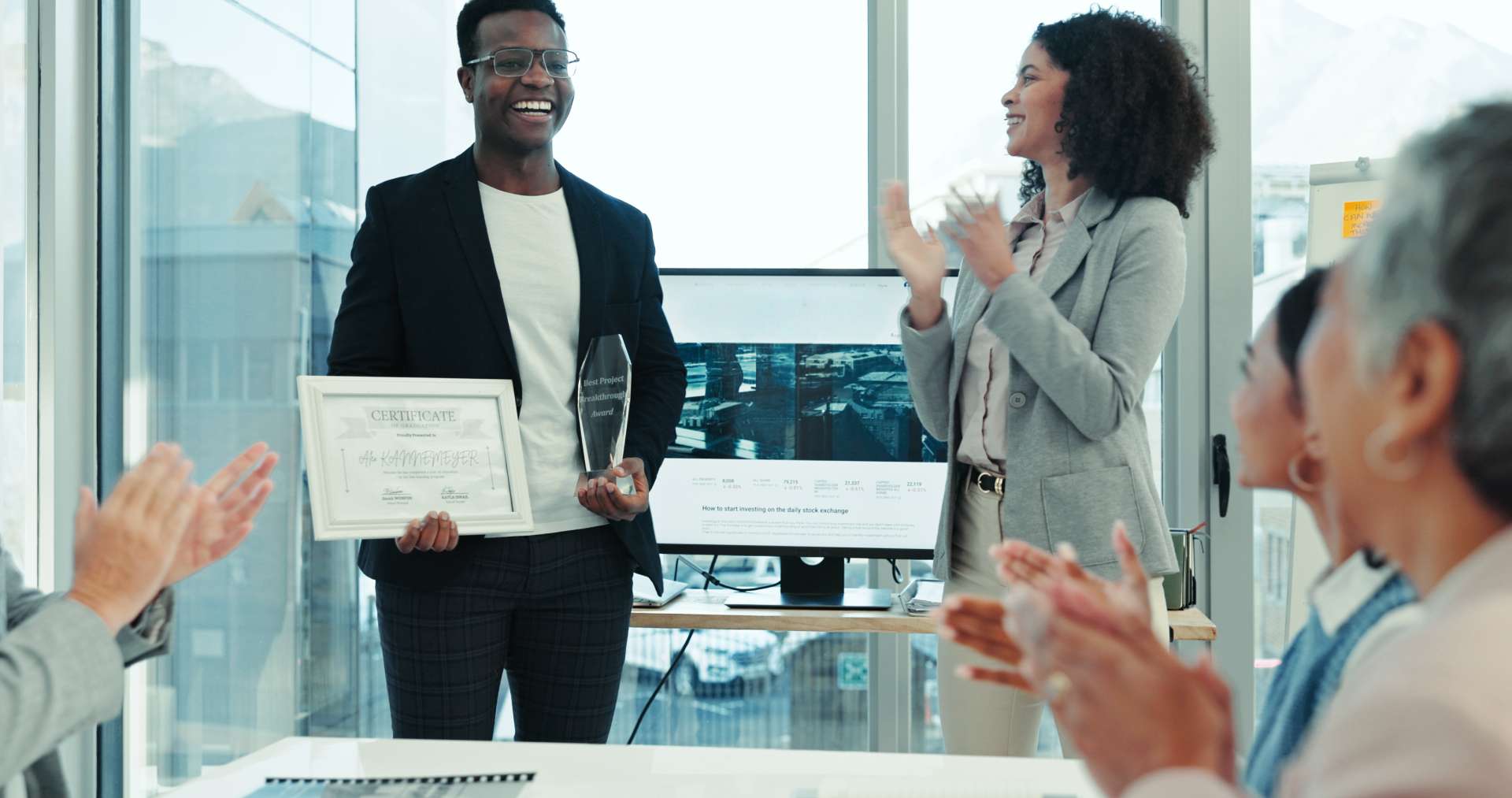 Person standing and holding a certificate and a glass award in a bright office setting, while others seated around a table applaud. A large window and a monitor are visible in the background.