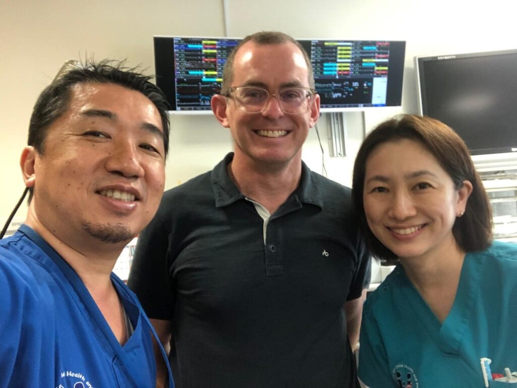A classroom-style setting with several people seated at wooden tables facing a projector screen. The screen displays a presentation slide with a diagram and text in yellow and red. A presenter stands near the screen, and the room includes ceiling-mounted projectors, fluorescent lighting, and a mix of chairs and tables arranged for a lecture.