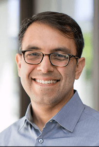 Person wearing a gray collared shirt in an indoor setting with blurred background.
