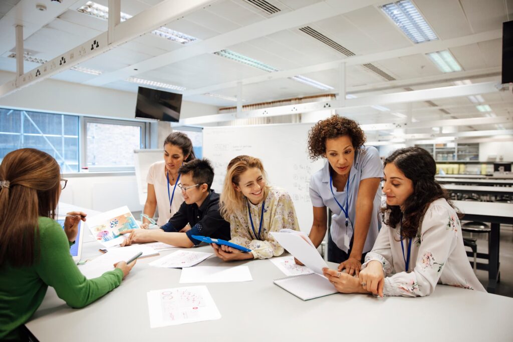 Group of six people gathered around a table in a bright classroom or office setting, collaborating on a project. They are reviewing papers, notebooks, and a tablet device. The background includes large windows, a whiteboard with writing, and overhead fluorescent lighting.