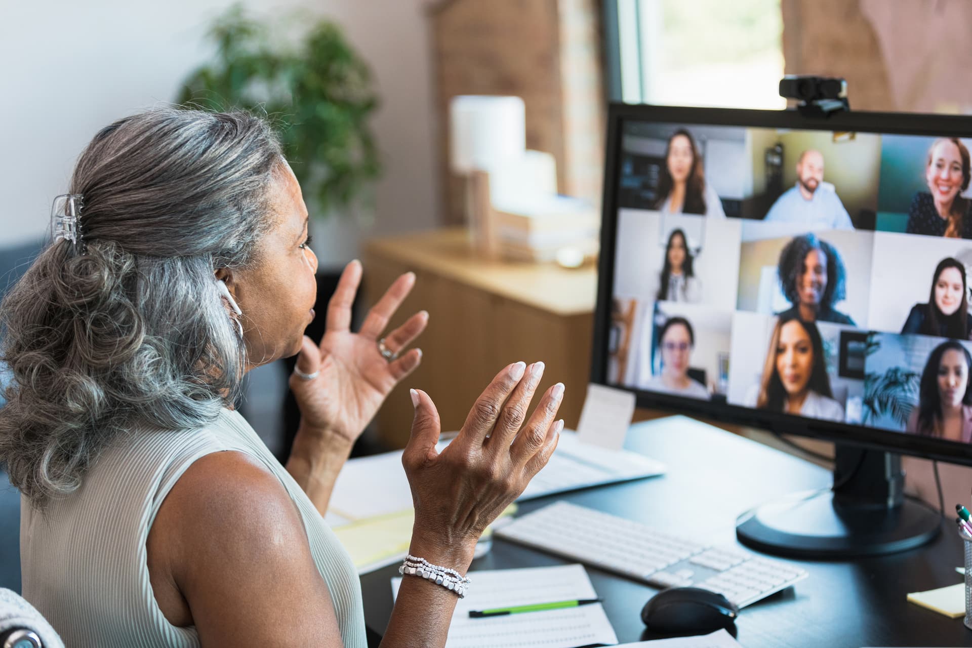Person sitting at a desk in front of a computer monitor displaying a video conference with multiple participants in a grid layout. The desk has a keyboard, mouse, notepad, and other office items, and the background shows a bright room with natural light.