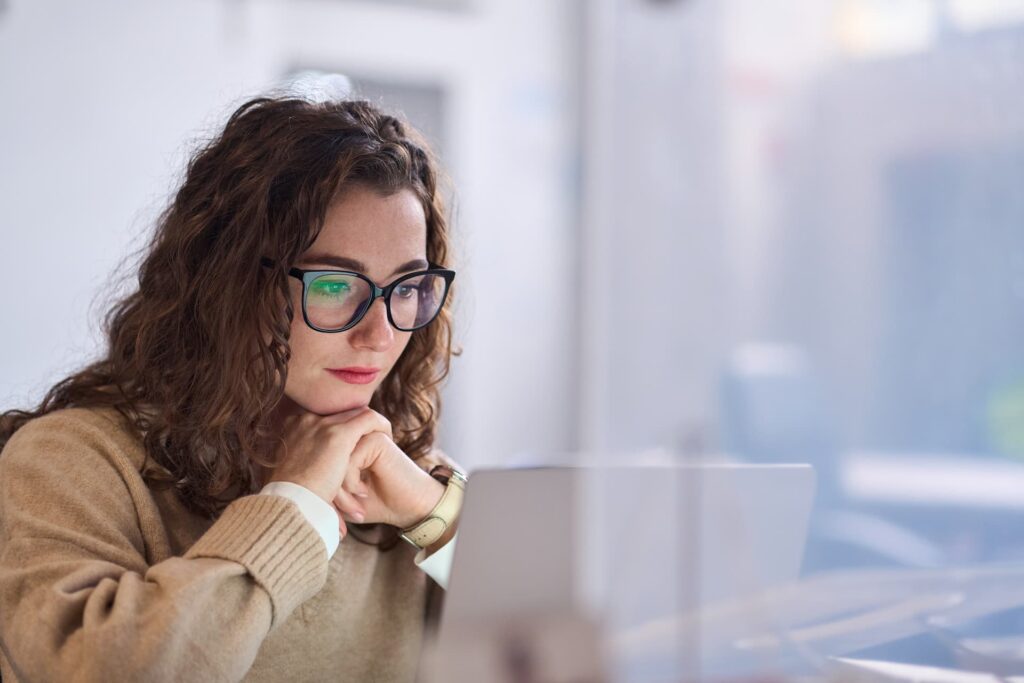 Person sitting at a desk in front of a laptop, wearing a light-colored sweater and glasses, with hands resting near the chin. The setting appears to be an office or workspace with a glass partition in the background.