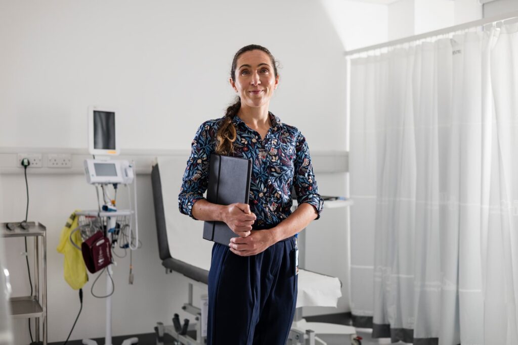 Person standing in a medical examination room holding a black folder. The room includes an exam chair, medical equipment on a stand, a monitor, and a privacy curtain in the background.