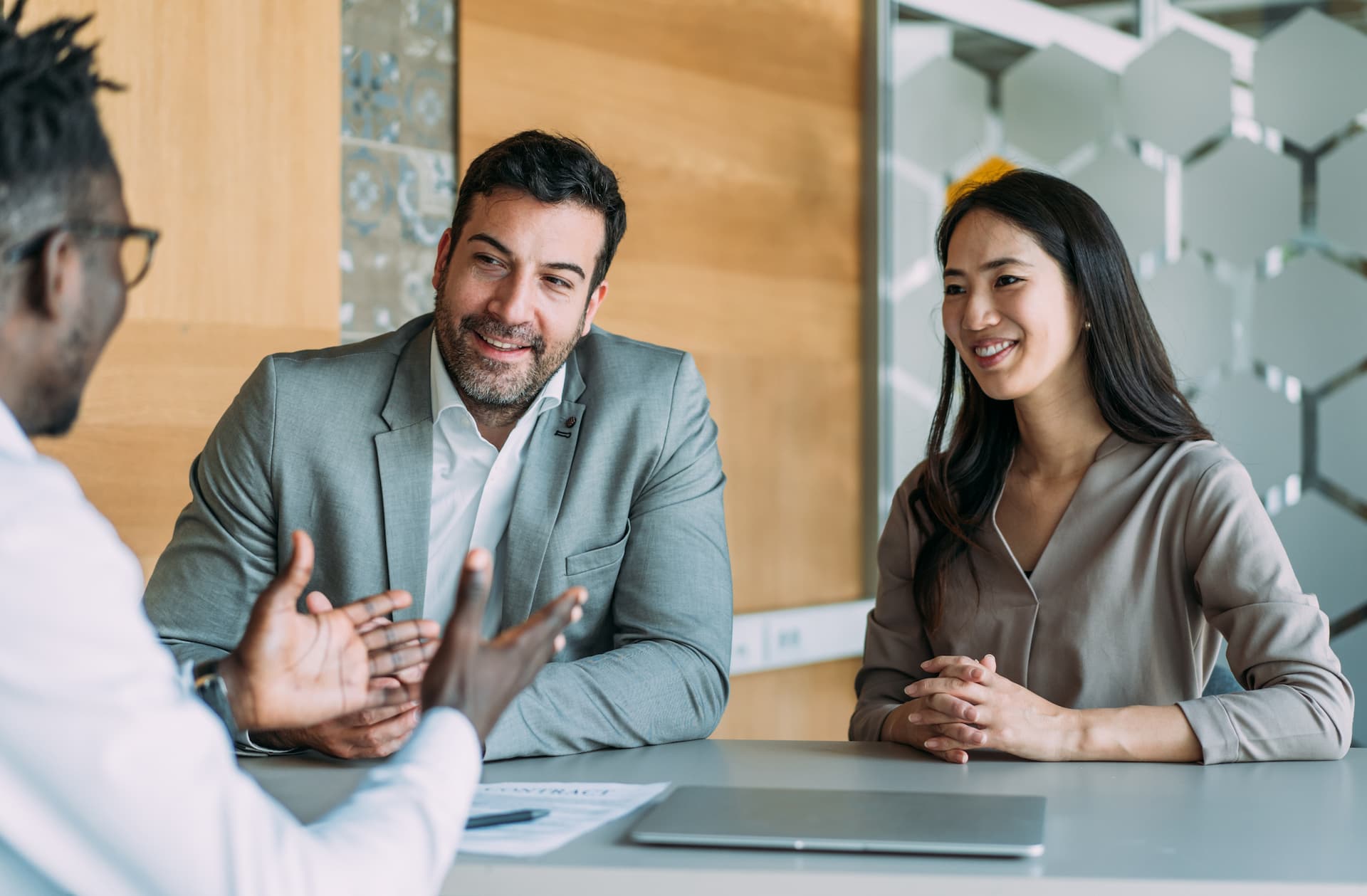 Three people seated at a table in a modern office setting, engaged in a discussion. One person is gesturing with hands while speaking, and two others are listening with hands resting on the table. A laptop and a document are visible on the table, and the background features wood paneling and decorative glass partitions.