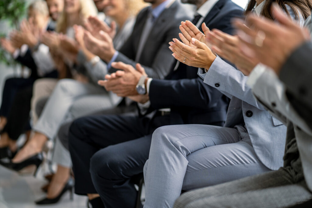 In a corporate setting, an audience of professionals energetically applauds a presentation, highlighting engagement, enthusiasm, and a thriving corporate culture.