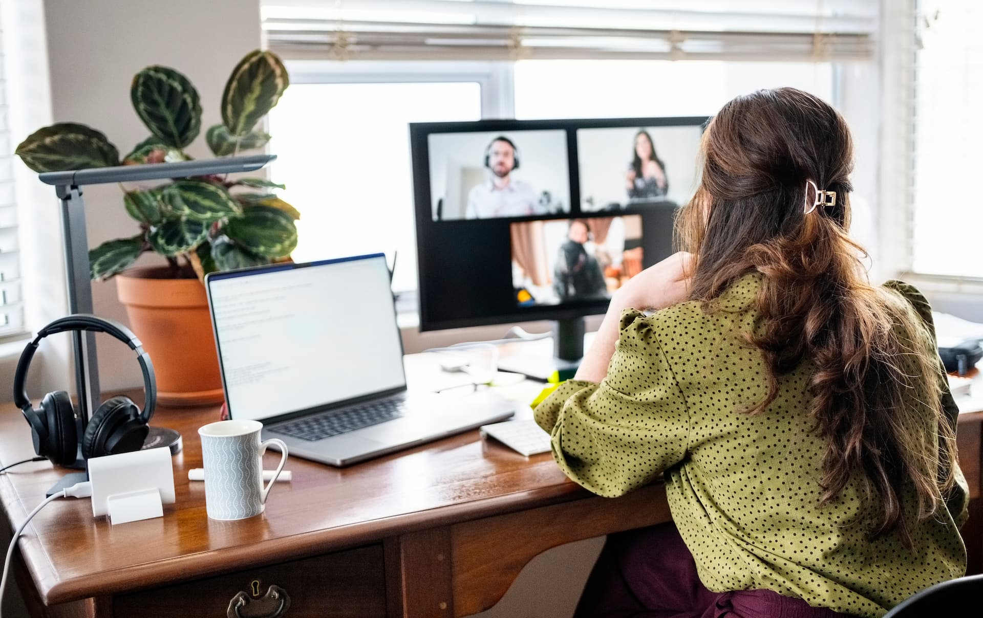 Person sitting at a wooden desk participating in a video conference on a monitor with four visible participants. The desk has a laptop displaying text, a coffee mug, headphones, a plant in a terracotta pot, and a desk lamp. Sunlight streams through blinds in the background.
