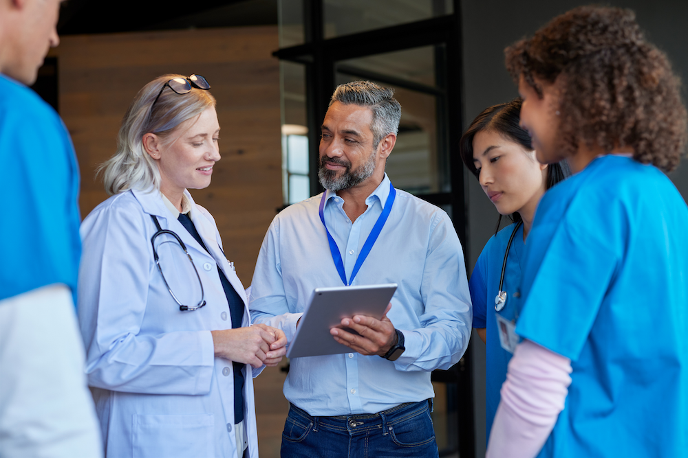 Healthcare professionals in medical attire stand together in discussion, with one person holding a tablet.
