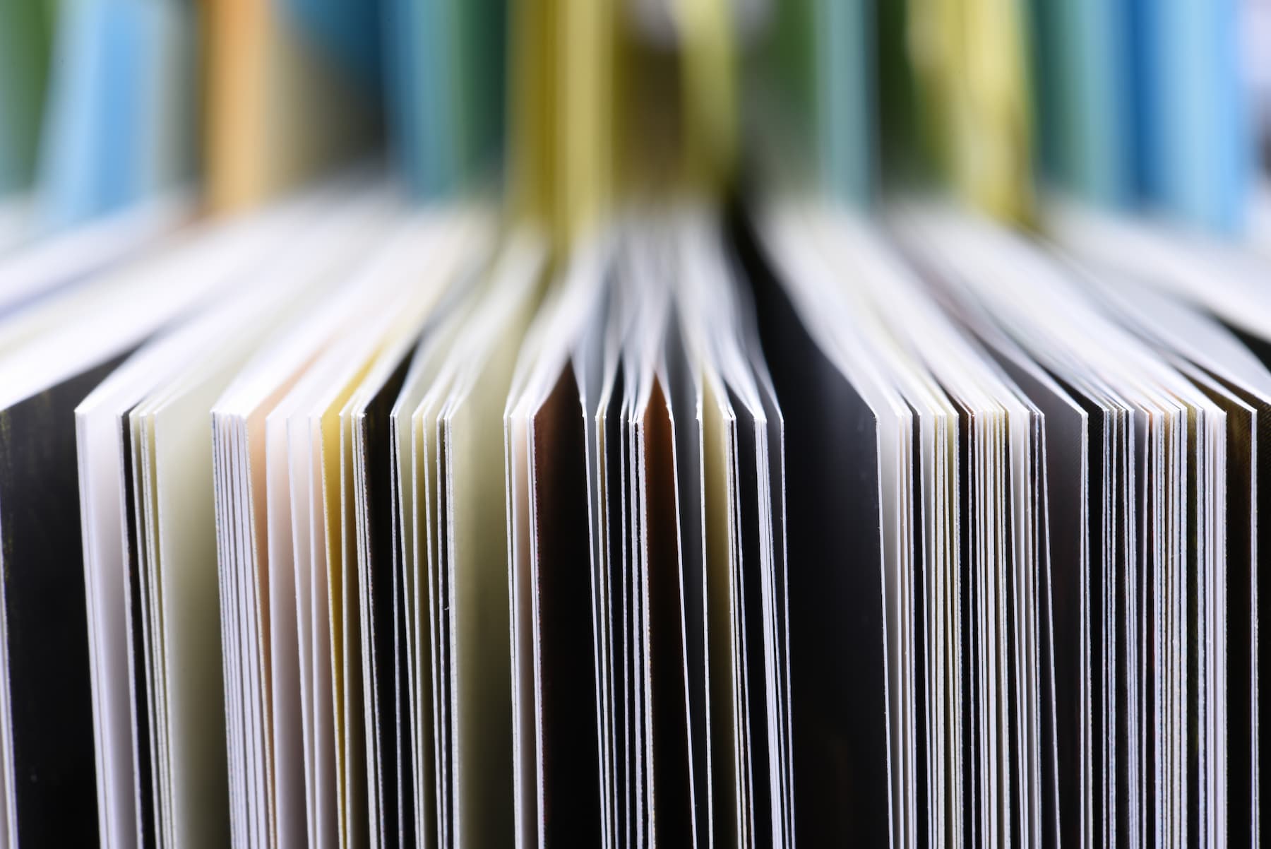 Close-up view of the edges of multiple open books or magazines arranged vertically, showing colorful pages and spines in soft focus.