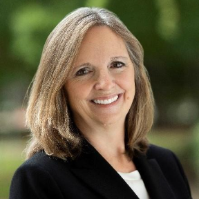 Person with shoulder-length light brown hair wearing a black blazer over a white top, smiling outdoors in front of a blurred green background