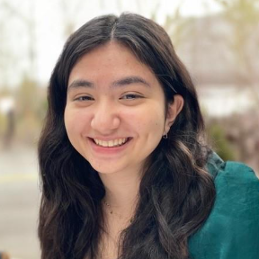 Person with long dark wavy hair wearing a dark green top and small hoop earrings, smiling at the camera in an outdoor setting with blurred trees and buildings in the background.