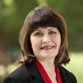 Person with shoulder-length brown hair wearing a black blazer over a red top, looking at the camera with a neutral expression in an outdoor setting with greenery in the background.