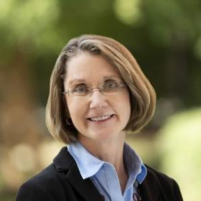 Person with short light brown hair wearing glasses, a light blue collared shirt, and a dark blazer, smiling at the camera in an outdoor setting with a blurred background of green and brown tones.