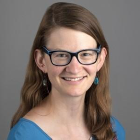 Person with long brown hair wearing blue-framed glasses, a blue top, and dangling earrings, smiling at the camera against a plain gray background.