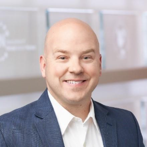Bald man wearing a blue textured suit jacket over a white dress shirt, smiling at the camera in an indoor setting with a softly blurred background featuring plaques and a circular emblem labeled 'International Research Center.