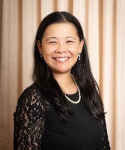 Person wearing a black dress with lace sleeves and accessorized with a pearl necklace and earrings, posed in front of a light-colored curtain backdrop.