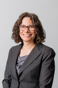 Person wearing a dark suit jacket over a gray draped top, posed against a plain light gray background.