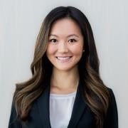 A person with long wavy hair, wearing a dark blazer and light top, poses against a plain light background.