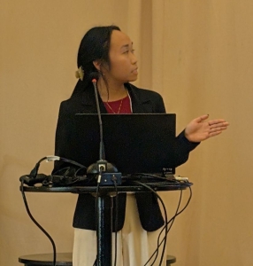 A person standing behind a podium with a laptop and microphone, gesturing with one hand while presenting in a conference or meeting setting. The background is plain, and several cables are visible on the podium.
