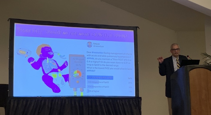 A person standing at a podium giving a presentation in a conference room. The large screen behind shows a colorful slide featuring an illustration of an infant connected to an oxygen device, along with text discussing “Floor FiO₂” and oxygen management in PPHN. The slide includes a poll with percentage results for different FiO₂ levels.
